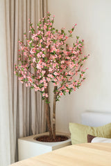 Die künstliche Kirschblütenpflanze 120 cm rosa mit ihren rosafarbenen Kirschblüten steht neben einem Holztisch in einem hellen, modernen Raum, dahinter ein beiger Vorhang und ein helles Sofa.