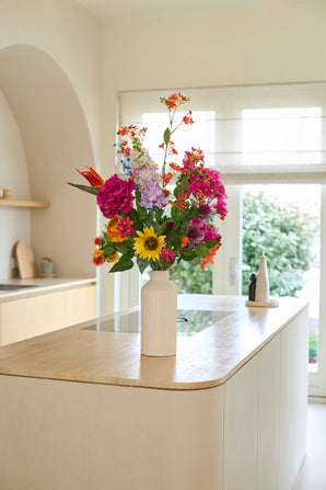 Eine weiße Vase mit dem Strauß „Colorful Spirit“ aus künstlichen Seidenblumen steht auf einer Kücheninsel aus hellem Holz in einer hellen, modernen Küche mit großen Fenstern und neutralem Dekor und schafft einen nachhaltigen und lebendigen Mittelpunkt.