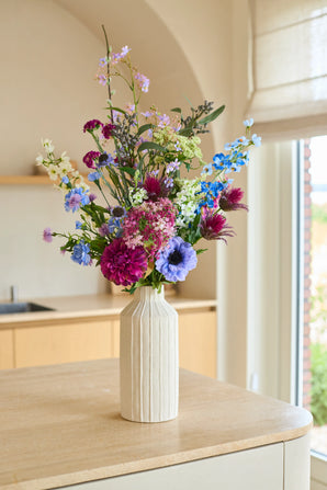 Der künstliche Blumenstrauß Purple Rain aus Seide, ein Arrangement aus violetten, blauen, rosa und weißen Seidenblumen, wird in einer hohen, cremefarbenen Keramikvase platziert. Dieser handgefertigte Blumenstrauß liegt anmutig auf einer hellen Holzarbeitsplatte und ist in sanftes, natürliches Licht getaucht – ein wahres Spiegelbild niederländischer Handwerkskunst.