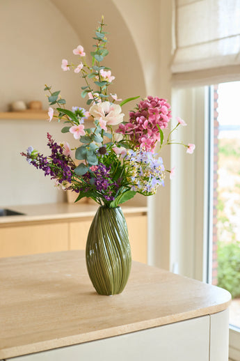 Auf einer hellen Kücheninsel in der Nähe eines Fensters, durch das natürliches Licht einfällt, steht eine grüne Vase mit rosa, lila und weißen Blumen aus dem Seiden-Kunstblumenstrauß „Lavender Dreams“.