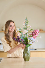 Eine Frau mit langen, welligen Haaren arrangiert einen Strauß „Lavender Dreams“ aus künstlichen Seidenblumen in einer grünen Vase in ihrer modernen Küche und lächelt, während sie arbeitet. Der lebendige Strauß aus Seidenblumen verleiht ihrem Zuhause zeitlose Schönheit.