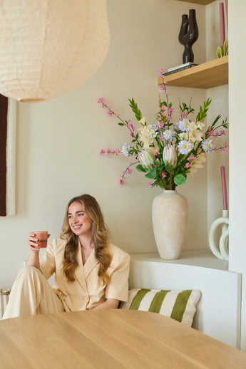 Eine Frau im cremefarbenen Anzug sitzt mit einer Tasse in der Hand und einem Lächeln im Gesicht auf einer Bank. Hinter ihr steht eine große Vase mit dem künstlichen Seidenblumenstrauß „Lots of Love XL“, bestehend aus rosa, weißen und gelben Blumen, mit Regalen im Hintergrund, auf denen Deko-Objekte ausgestellt sind. Das Ambiente wirkt gemütlich und modern.