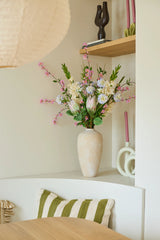Auf einem weißen Regal steht eine beige Vase mit dem bunten Strauß künstlicher Seidenblumen „Lots of Love XL“, daneben rosa Kerzen und Deko-Objekte. Auf einem Stuhl unter dem Regal liegt ein grün-weiß gestreiftes Kissen.