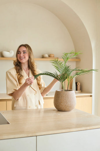 Eine Frau mit langen blonden Haaren lächelt, während sie in einer modernen, in neutralen Farbtönen gehaltenen Küche mit minimalistischem Dekor eine 18 Zoll große künstliche Chamaedorea-Palme im Topf pflegt.