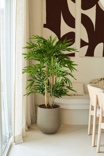 Eine künstliche Bambuspflanze von 120 cm steht in einem modernen, hellen Wohnzimmer an einem Fenster mit Gardinen, neben einem weißen Stuhl und abstrakter Wandkunst.