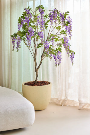 Die künstliche Wisteria-Pflanze (210 cm) mit ihren überhängenden violetten Blütensträußen wird im Innenbereich neben durchscheinenden beigen Vorhängen und einem hellen Sofa platziert.