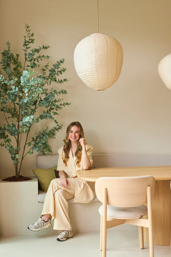 Eine Frau in einem beigen Anzug sitzt lächelnd an einem Holztisch in einem modernen, minimalistischen Raum mit hellen Wänden, von der Decke hängenden Papierpendellampen und einer 180 cm großen künstlichen Eukalyptuspflanze, die dem Raum etwas Grün verleiht.