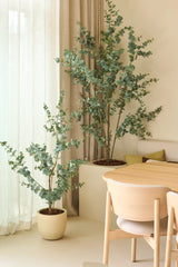 Zwei künstliche Eukalyptus-Kunstpflanzen von 150 cm, platziert neben transparenten Vorhängen und beigen Wänden, kombiniert mit einem Esstisch und Stühlen aus hellem Holz in einem minimalistischen, modernen Interieur.