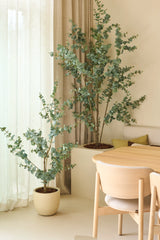 Zwei künstliche Eukalyptus-Kunstpflanzen von 150 cm, platziert neben transparenten Vorhängen und beigen Wänden, kombiniert mit einem Esstisch und Stühlen aus hellem Holz in einem minimalistischen, modernen Interieur.