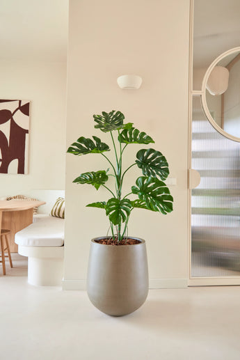 Die 120 cm große Kunstpflanze Monstera wird in einem modernen, minimalistischen Wohnraum mit neutralen Wänden, einem runden Spiegel, Holzmöbeln und abstrakter Wandkunst platziert.