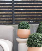 Auf einer Terrasse neben einem beigen Gartenstuhl, vor einem modernen schwarzen Lattenzaun und kahlen Bäumen stehen drei künstliche Buchsbaum-Rundformschnitte im römischen UV-Stil (D30 cm).