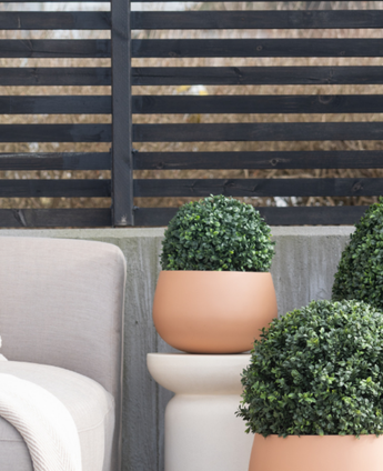Auf einer Terrasse neben einem beigen Gartenstuhl, vor einem modernen schwarzen Lattenzaun und kahlen Bäumen stehen drei künstliche Buchsbaum-Rundformschnitte im römischen UV-Stil (D30 cm).