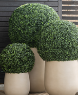 Drei große Kunstpflanzen-Buchsbaum-UV-Töpfe im römischen Stil (D65 cm) präsentieren ein sauber beschnittenes, kugelförmiges grünes Formgehölz vor einem Hintergrund aus dunklen Holzlatten auf einem Holzboden.
