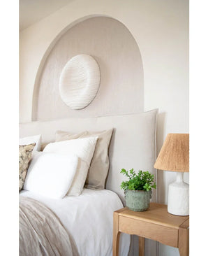 Ein gemütliches Schlafzimmer mit beigem Kopfteil, neutraler Bettwäsche und Kissen, einem hölzernen Nachttisch mit der 27 cm großen künstlichen Eukalyptuspflanze, einer Lampe mit strukturiertem Schirm und runder weißer Wandkunst über dem Bett in einer gewölbten Nische.