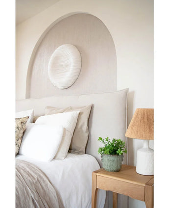Ein gemütliches Schlafzimmer mit beigem Kopfteil, neutraler Bettwäsche und Kissen, einem hölzernen Nachttisch mit der 27 cm großen künstlichen Eukalyptuspflanze, einer Lampe mit strukturiertem Schirm und runder weißer Wandkunst über dem Bett in einer gewölbten Nische.