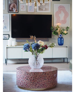 Ein modernes Wohnzimmer verfügt über einen Terrazzo-Couchtisch mit einer Glasvase mit Blumen, einen Fernseher an der Wand, gerahmte Kunst und Fotos, ein weißes Sideboard und die künstliche Hortensie Deluxe 57 cm lila als elegantes blaues Blumenarrangement im Hintergrund.