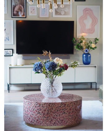 Ein modernes Wohnzimmer verfügt über einen Terrazzo-Couchtisch mit einer Glasvase mit Blumen, einen Fernseher an der Wand, gerahmte Kunst und Fotos, ein weißes Sideboard und die künstliche Hortensie Deluxe 57 cm lila als elegantes blaues Blumenarrangement im Hintergrund.