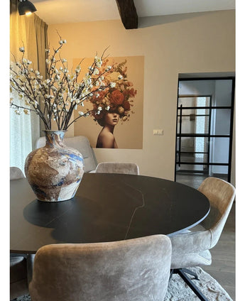 Ein moderner Essbereich mit einem runden schwarzen Tisch, beigen Stühlen und einer großen weißen Keramikvase mit künstlichen japanischen Kirschblüten (94 cm). Den Hintergrund schmückt ein Wandgemälde einer Frau mit Blumen im Haar.