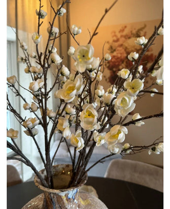Eine Nahaufnahme der künstlichen japanischen Kirschblüte 94 cm mit anmutigen weißen künstlichen Kirschblüten mit gelben Zentren auf braunen Zweigen in einer Glasvase, auf einem dunklen Tisch mit einem eleganten, sanft beleuchteten Raum im Hintergrund.