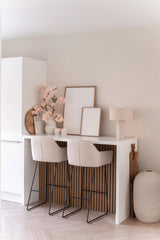 Eine minimalistische Hausbar mit zwei beigen Hockern mit Kissen, einer weißen Theke, gerahmter Kunst, einer Tischlampe, neutraler Dekoration und künstlichen Kirschblüten (100 cm) in Hellrosa in Vasen vor einer hellen Wand.