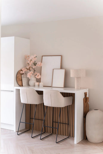 Eine minimalistische Hausbar mit zwei beigen Hockern mit Kissen, einer weißen Theke, gerahmter Kunst, einer Tischlampe, neutraler Dekoration und künstlichen Kirschblüten (100 cm) in Hellrosa in Vasen vor einer hellen Wand.
