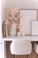 Eine minimalistische Schreibtischanordnung mit einem weißen Stuhl, zwei Kerzen, dekorativen Tabletts, gerahmter abstrakter Kunst und einer 100 cm großen künstlichen Kirschblüte in Hellrosa in einer weißen Vase auf einer weißen Oberfläche.