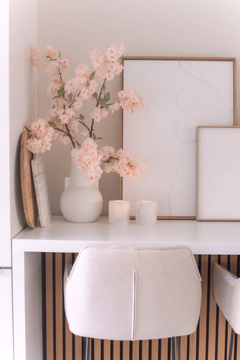 Eine minimalistische Schreibtischanordnung mit einem weißen Stuhl, zwei Kerzen, dekorativen Tabletts, gerahmter abstrakter Kunst und einer 100 cm großen künstlichen Kirschblüte in Hellrosa in einer weißen Vase auf einer weißen Oberfläche.