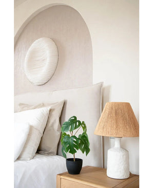 Ein ordentlich gemachtes Bett mit weißer Bettwäsche steht neben einem hölzernen Nachttisch mit der Art Monstera 40 cm in einem schwarzen Dekotopf und einer weißen Lampe mit geflochtenem Schirm. An der Wand hängen ein weicher Bogen und ein rundes, strukturiertes Kunstwerk.