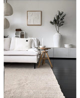 Ein minimalistisches Wohnzimmer mit einem hellen Sofa, einem neutralen Teppich, einem weißen Sideboard, einem künstlichen Olivenbaum 120 cm als Dekoelement, abstrakter Wandkunst und Holzhockern – alles in einer sanften mediterranen Atmosphäre mit neutralen Tönen.