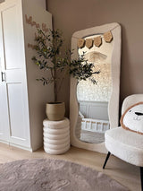 Ein gemütliches, modernes Kinderzimmer in neutralen Farbtönen verfügt über einen gewellten Bodenspiegel, den künstlichen Olivenbaum 120 cm für einen mediterranen Touch, einen Plüschstuhl mit Bärengesichtskissen und ein im Spiegel reflektiertes Kinderbett – die perfekte Dekoration für jeden Raum.
