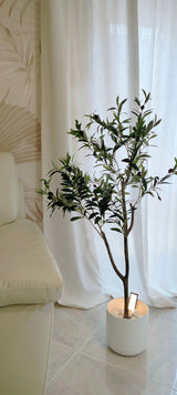 Ein künstlicher Olivenbaum 120 cm mit grünen Blättern steht auf einem Fliesenboden neben einem cremefarbenen Sofa und verleiht dem Raum eine mediterrane Atmosphäre, vor weißen Vorhängen und einer Wand mit dezentem Muster.