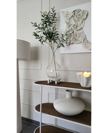 Ein minimalistischer Konsolentisch zeigt eine klare Vase mit dem künstlichen Olivenzweig 109 cm, eine runde weiße Keramikvase und eine weiße Schale mit LED-Kerzen. Darüber hängt strukturierte weiße Wandkunst und daneben steht eine Stehlampe.