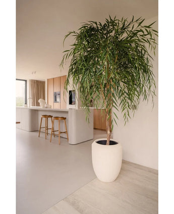 Ein künstlicher Weidenbaum 210 cm mit langen, schlanken Blättern bringt Grün in eine moderne Küche mit hellen Holzschränken, einer weißen Insel und Barhockern aus Holz. Sonnenlicht, das durch das Fenster fällt, unterstreicht seine Präsenz im minimalistischen Raum.