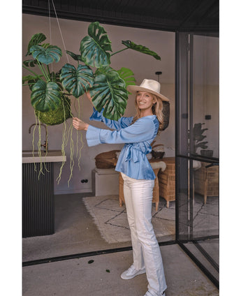 Eine lächelnde Frau mit Hut, blauer Bluse und weißer Hose steht drinnen und hält eine künstliche Hängepflanze Monstera Deluxe 60 cm mit großen grünen Blättern, umgeben von modernem Dekor für eine tropische Atmosphäre mit Dekopflanzen.