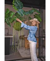 Eine lächelnde Frau in blauer Bluse, weißer Hose und Hut zeigt die künstliche Hängepflanze Monstera Deluxe 80 cm mit gewundenen grünen Ranken, die mit Glaswänden und Rattanmöbeln eine tropische Atmosphäre in den hellen Innenraum bringt.