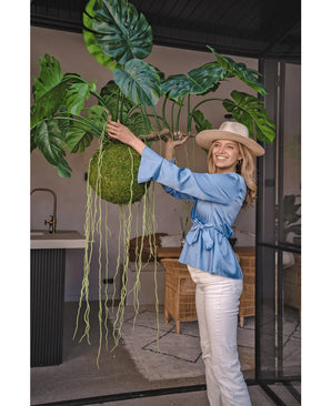 Eine lächelnde Frau in blauer Bluse, weißer Hose und Hut zeigt die künstliche Hängepflanze Monstera Deluxe 80 cm mit gewundenen grünen Ranken, die mit Glaswänden und Rattanmöbeln eine tropische Atmosphäre in den hellen Innenraum bringt.