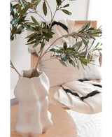 Eine weiße, geformte Vase hält Art-Olivenblatt 90 cm UV mit grünen Blättern auf einem hellen Holztisch. Dahinter verstärken ein beiger Stuhl und zwei cremefarbene Kissen mit auffälligen schwarzen Streifen die mediterrane Atmosphäre.