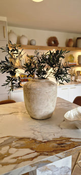 Eine rustikale beige Vase mit feuerfestem künstlichem Olivenblatt (90 cm) steht auf einer Marmorarbeitsplatte in einer modernen, warm beleuchteten Küche mit Regalen und mediterraner Dekoration im Hintergrund.