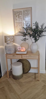 Ein hölzerner Konsolentisch mit zwei Schubladen enthält eine Lampe, eine große weiße Vase mit künstlichem Olivenblatt (90 cm, feuerfest), eine kleinere Vase, Bücher und Dekosteine. Mediterrane Akzente und gerahmte abstrakte Kunst runden den Look oben ab. Unten stehen ein Hocker und ein rundes Tablett.