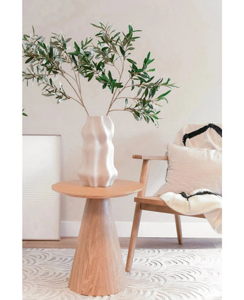 In einem modernen, minimalistischen Wohnraum mit mediterranem Dekor steht eine geformte weiße Vase mit dem feuerhemmenden künstlichen Olivenblatt 90 cm auf einem runden Holztisch neben einem hellen Holzstuhl mit weißen und schwarzen Kissen.