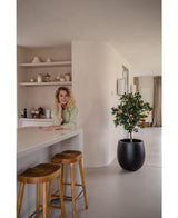 Eine Frau mit blonden Locken lächelt und lehnt an einer Theke. Neben ihr sorgen zwei Holzhocker und ein künstlicher Orangenbaum 120 cm für eine frische Atmosphäre in der modernen, hellen Küche mit Regalen und Tellern dahinter.