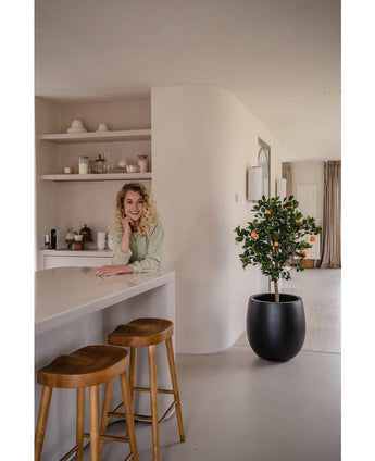 Eine Frau mit blonden Locken lächelt und lehnt an einer Theke. Neben ihr sorgen zwei Holzhocker und ein künstlicher Orangenbaum 120 cm für eine frische Atmosphäre in der modernen, hellen Küche mit Regalen und Tellern dahinter.