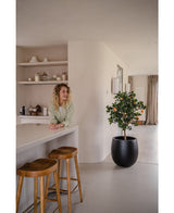 Eine Frau mit blonden Locken lächelt hinter einer Theke mit zwei Holzhockern. Der künstliche Orangenbaum 120 cm im Topf verleiht dem modernen, minimalistischen Raum, der in neutralen Farbtönen dekoriert ist, Charme.