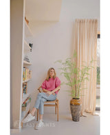 Eine Frau in rosa Pullover und Jeans sitzt auf einem Holzstuhl neben einer 120 cm großen künstlichen Farnpflanze, mit Bücherregalen und einem Fenster mit beigen Vorhängen in einem hellen, modernen Raum.