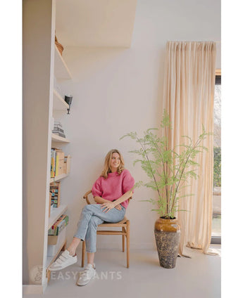 Eine Frau in rosa Pullover und Jeans sitzt auf einem Holzstuhl neben einer 120 cm großen künstlichen Farnpflanze, mit Bücherregalen und einem Fenster mit beigen Vorhängen in einem hellen, modernen Raum.