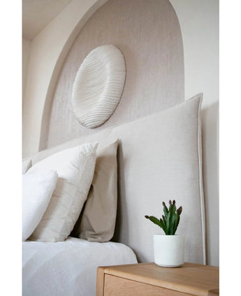 Ein modernes Schlafzimmer verfügt über ein beige gepolstertes Kopfteil, weiße Kissen, eine 20 cm große künstliche Sukkulente auf einem Nachttisch aus Holz und runde, strukturierte Wandkunst über dem Bett.
