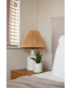 Ein Nachttisch mit einer weißen Lampe mit geflochtenem beigem Schirm und der künstlichen Sukkulente 20 cm steht neben einem Bett mit weiß-beiger Bettwäsche, in der Nähe eines Fensters mit Holzjalousien.