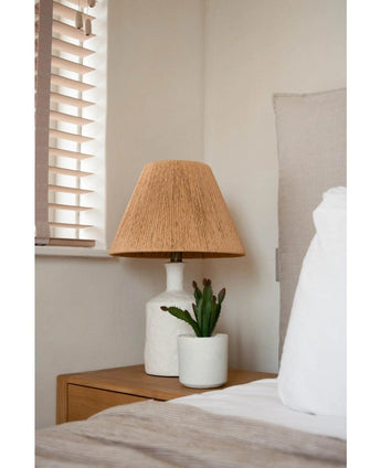 Ein Nachttisch mit einer weißen Lampe mit geflochtenem beigem Schirm und der künstlichen Sukkulente 20 cm steht neben einem Bett mit weiß-beiger Bettwäsche, in der Nähe eines Fensters mit Holzjalousien.