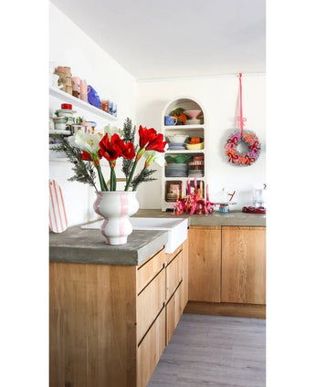 Eine helle Küche mit Holzschränken, offenen weißen Regalen mit Tellern und Töpfen, einem festlichen Kranz an der Wand und einer künstlichen Blume Amaryllis Real Touch rot 36 cm auf der Betonarbeitsplatte.