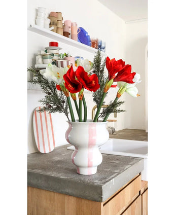 Die Kunstblume Amaryllis Real Touch rot 36 cm steht in einer weiß-rosa gestreiften Keramikvase auf einer Betonarbeitsplatte in einer hellen Küche mit Regalen und buntem Geschirr im Hintergrund.